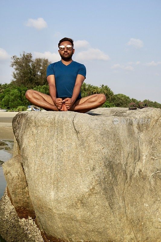 印度男子在岩石上练习蝴蝶/鞋匠姿势(Baddha Konasana)瑜伽姿势，水边，Palolem海滩，果阿，印度